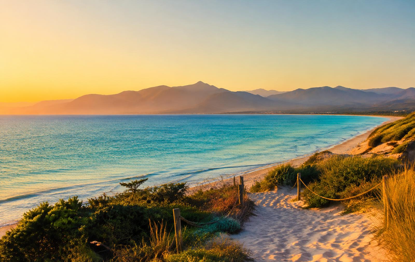 Sandy path leading down to a turquoise bay with mountains at sunset